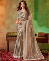 A model wearing a pastel brown fancy satin swarovski and stud work saree with cut work border, standing in front of a green backdrop with rose petals scattered around.